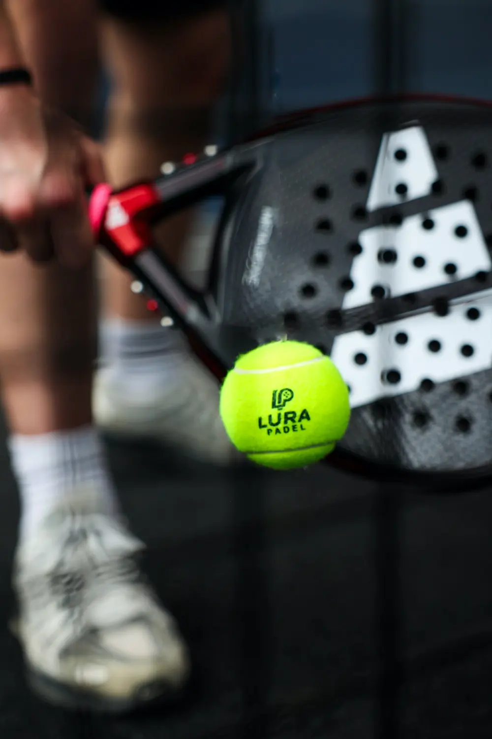 A LURA Padel ball at the moment it meets the racket face, a player blurred behind.